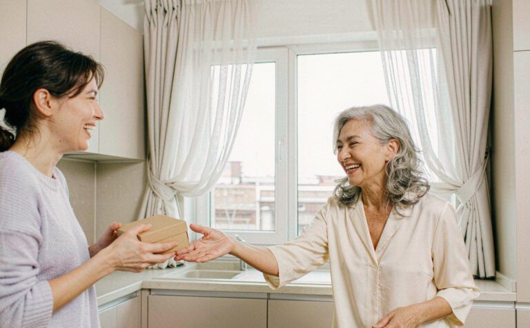 A daughter handing a gift box to her mother across a kitchen counter, both laughing